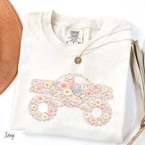 Op de afbeelding: Ivoorkleurig T-shirt met een bloemenmonster truck ontwerp. De truck is gevuld met roze, gele en blauwe bloemen. Het shirt heeft een ronde hals en korte mouwen. Een gouden ketting met een hanger is over het shirt gedrapeerd.