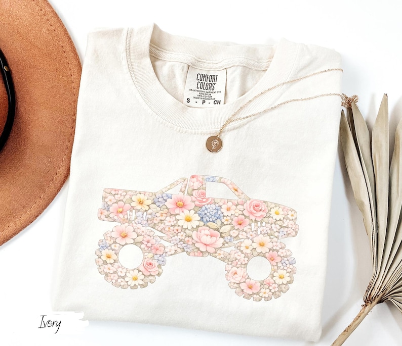 Op de afbeelding: Ivoorkleurig T-shirt met een bloemenmonster truck ontwerp. De truck is gevuld met roze, gele en blauwe bloemen. Het shirt heeft een ronde hals en korte mouwen. Een gouden ketting met een hanger is over het shirt gedrapeerd.
