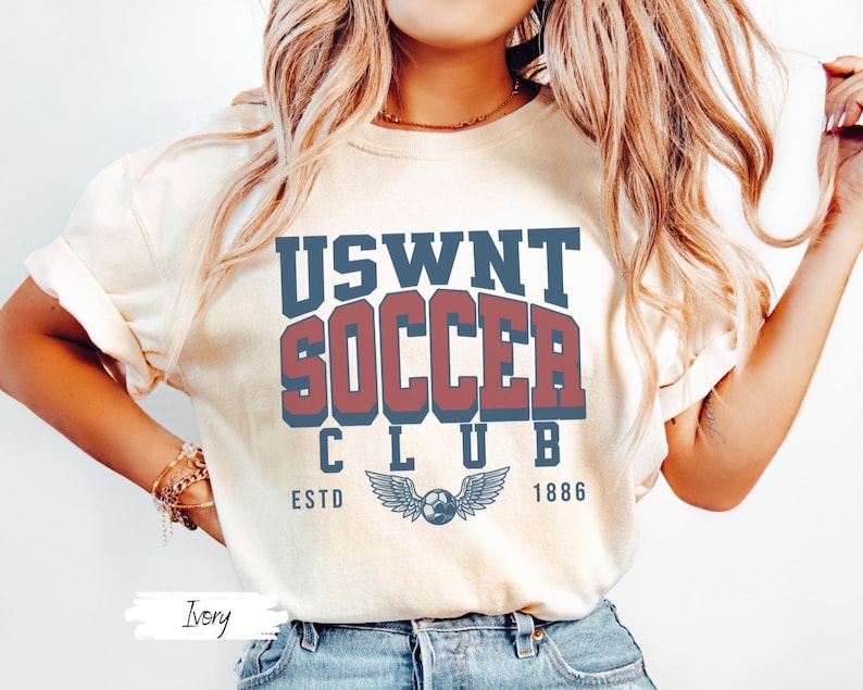 Puede incluir: Una camiseta blanca con el texto "USWNT SOCCER CLUB ESTD 1886" en letras azules y rojas. La camiseta presenta un gr&aacute;fico de un bal&oacute;n de f&uacute;tbol con alas.