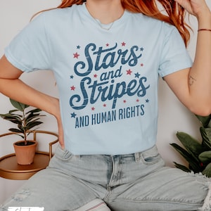 Puede incluir: Camiseta azul claro con el texto "Stars and Stripes AND HUMAN RIGHTS" en escritura azul marino y acentos de estrellas rojas. La camiseta es de cuello redondo y est&aacute; hecha de un material suave.
