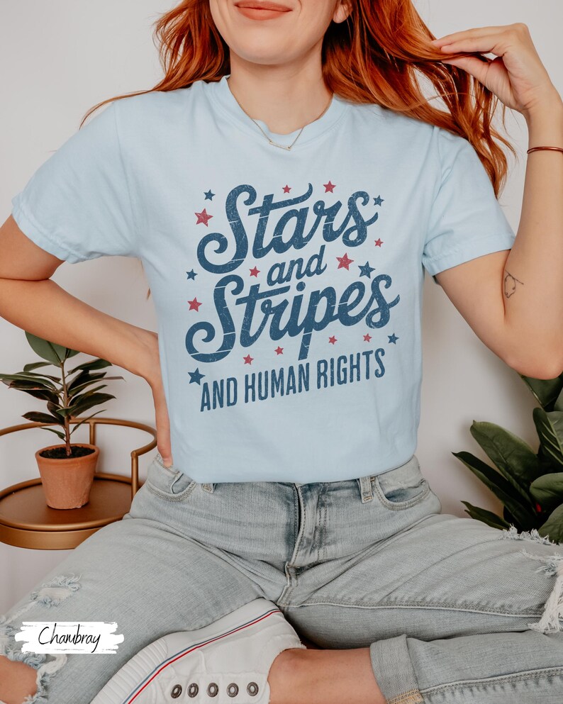 Puede incluir: Camiseta azul claro con el texto "Stars and Stripes AND HUMAN RIGHTS" en escritura azul marino y acentos de estrellas rojas. La camiseta es de cuello redondo y est&aacute; hecha de un material suave.