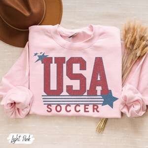May include: Light pink sweatshirt with a vintage-style graphic of "USA SOCCER" in red and blue with stars.