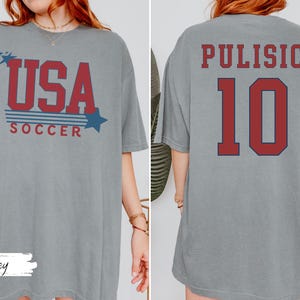 May include: A gray t-shirt with the text "USA SOCCER" in red and blue, with star graphics. The back of the shirt displays "PULISIC 10" in red. The shirt is a comfortable, casual style.