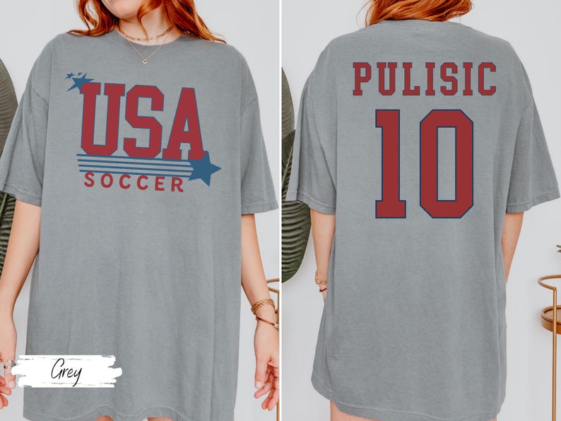 May include: A gray t-shirt with the text "USA SOCCER" in red and blue, with star graphics. The back of the shirt displays "PULISIC 10" in red. The shirt is a comfortable, casual style.