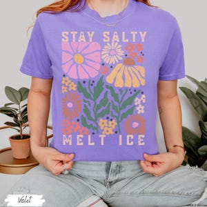 Op de afbeelding: Een violetkleurig T-shirt met de tekst "STAY SALTY MELT ICE" in een rechthoekig ontwerp. Het ontwerp bevat kleurrijke bloemen in roze, oranje en geel, met groene bladeren. Het shirt heeft korte mouwen.