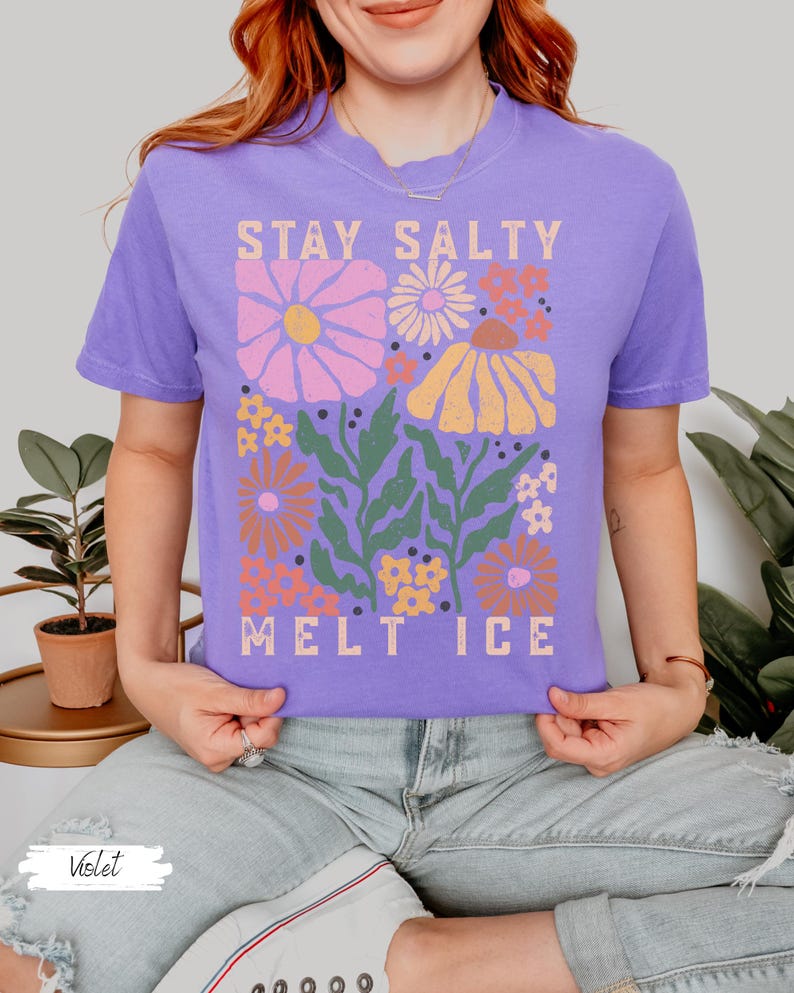 Op de afbeelding: Een violetkleurig T-shirt met de tekst "STAY SALTY MELT ICE" in een rechthoekig ontwerp. Het ontwerp bevat kleurrijke bloemen in roze, oranje en geel, met groene bladeren. Het shirt heeft korte mouwen.
