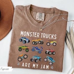 May include: Espresso-coloured t-shirt with the text "MONSTER TRUCKS ARE MY JAM" and colourful monster truck graphics. The shirt has a crew neck and short sleeves. A silver necklace with a pendant is draped across the shirt.