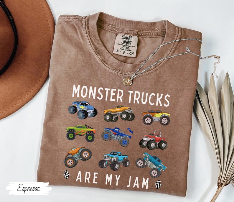 May include: Espresso-coloured t-shirt with the text "MONSTER TRUCKS ARE MY JAM" and colourful monster truck graphics. The shirt has a crew neck and short sleeves. A silver necklace with a pendant is draped across the shirt.