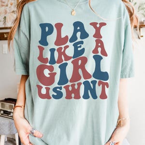 May include: A light blue t-shirt with the text "PLAY LIKE A GIRL USHNT" in a retro, colorful font.