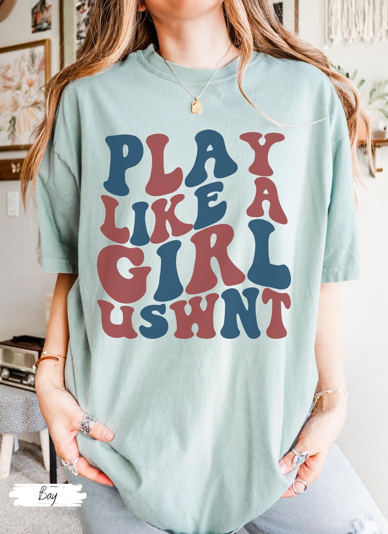 May include: A light blue t-shirt with the text "PLAY LIKE A GIRL USHNT" in a retro, colorful font.