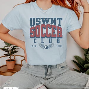 Puede incluir: Una camiseta azul claro con un gr&aacute;fico de estilo vintage que dice "USWNT Soccer Club EstD 1886" con un bal&oacute;n de f&uacute;tbol y alas.