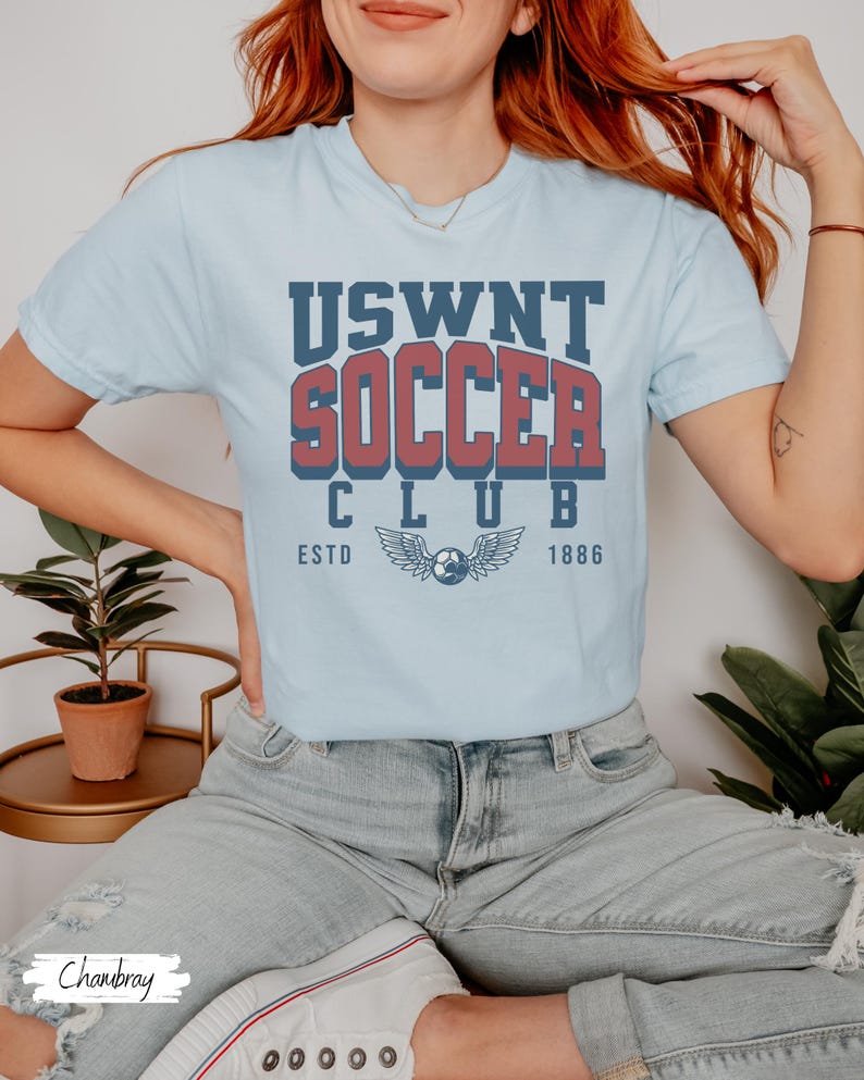Puede incluir: Una camiseta azul claro con un gr&aacute;fico de estilo vintage que dice "USWNT Soccer Club EstD 1886" con un bal&oacute;n de f&uacute;tbol y alas.