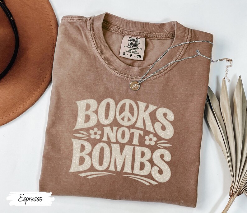May include: Espresso-colored t-shirt with the text "BOOKS NOT BOMBS" in a retro font. The shirt features a peace sign and flower graphic. A silver necklace with a pendant is draped on the shirt.