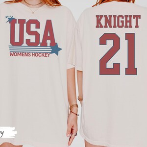 May include: Ivory-colored t-shirt with the text "USA WOMENS HOCKEY" in red and blue. The back of the shirt displays "KNIGHT 21" in the same colors. The shirt has a relaxed fit.