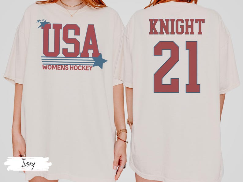 May include: Ivory-colored t-shirt with the text "USA WOMENS HOCKEY" in red and blue. The back of the shirt displays "KNIGHT 21" in the same colors. The shirt has a relaxed fit.