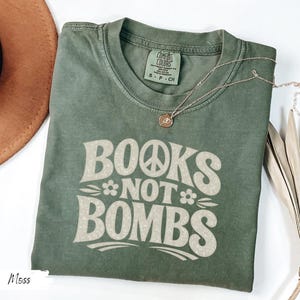 May include: Moss green t-shirt with the text "BOOKS NOT BOMBS" in a retro font. The shirt has a crew neck and short sleeves. A gold necklace with a pendant is draped on the shirt.