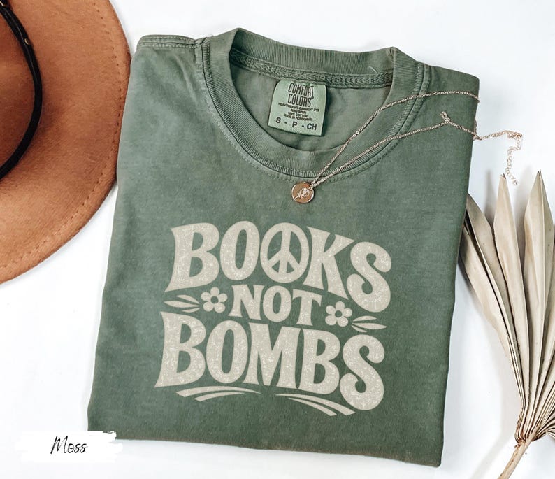 May include: Moss green t-shirt with the text "BOOKS NOT BOMBS" in a retro font. The shirt has a crew neck and short sleeves. A gold necklace with a pendant is draped on the shirt.