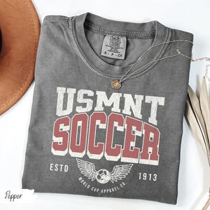 Może przedstawiać: Szara koszulka USMNT Soccer z postarzanym wzorem. Koszulka zawiera napis "USMNT SOCCER" w kolorze białym i czerwonym, a także piłkę nożną ze skrzydłami i napis "ESTD 1913". Złoty naszyjnik spoczywa na koszulce.