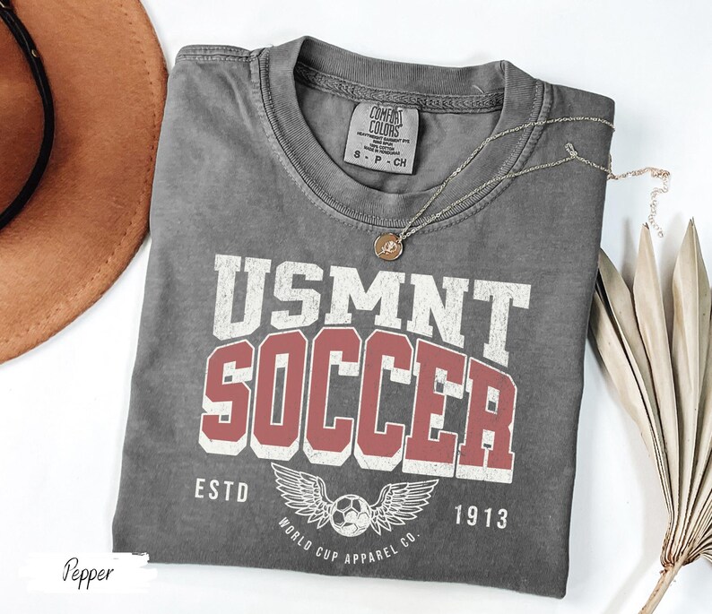Może przedstawiać: Szara koszulka USMNT Soccer z postarzanym wzorem. Koszulka zawiera napis "USMNT SOCCER" w kolorze białym i czerwonym, a także piłkę nożną ze skrzydłami i napis "ESTD 1913". Złoty naszyjnik spoczywa na koszulce.