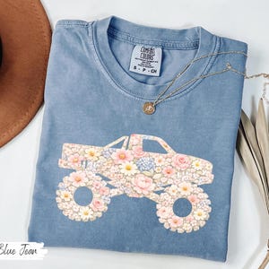 Op de afbeelding: Blauw spijkerbroekkleurig T-shirt met een bloemenmonster truck-afbeelding. De truck is gevuld met roze, witte en blauwe bloemen. Het shirt heeft een ronde hals en korte mouwen. De tekst "Blue Jean" staat linksonder.