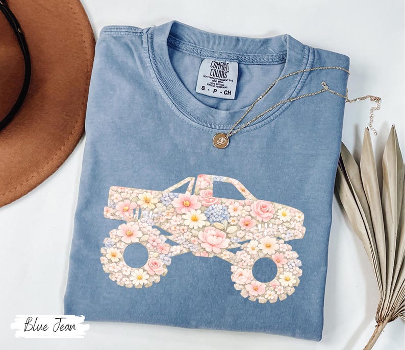 Op de afbeelding: Blauw spijkerbroekkleurig T-shirt met een bloemenmonster truck-afbeelding. De truck is gevuld met roze, witte en blauwe bloemen. Het shirt heeft een ronde hals en korte mouwen. De tekst "Blue Jean" staat linksonder.