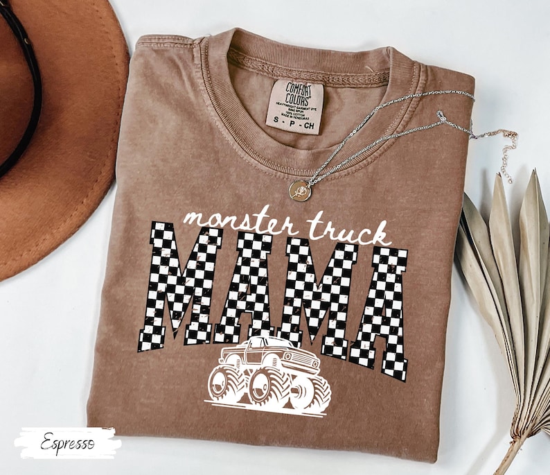 May include: Espresso-colored t-shirt with the words "monster truck MAMA" in white and a checkered pattern. A white monster truck graphic is below the text. The shirt has a crew neck and short sleeves. A silver necklace with a pendant is on the shirt.