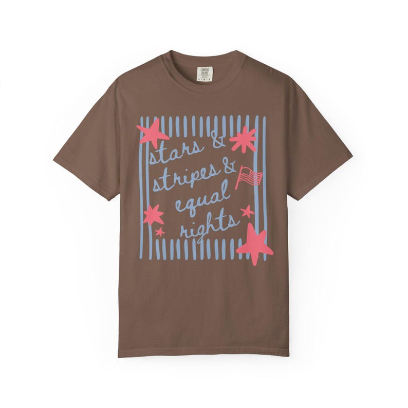 Puede incluir: Una camiseta marr&oacute;n con el texto "stars & stripes & equal rights" en un dise&ntilde;o azul y rosa. El dise&ntilde;o incluye estrellas, rayas y una peque&ntilde;a bandera americana. Una prenda informal para el d&iacute;a a d&iacute;a.