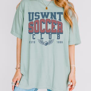 Puede incluir: Una camiseta de gran tama&ntilde;o de color verde claro con un dise&ntilde;o gr&aacute;fico de estilo vintage que dice "USWNT SOCCER CLUB ESTD 1886". El gr&aacute;fico presenta un bal&oacute;n de f&uacute;tbol con alas.
