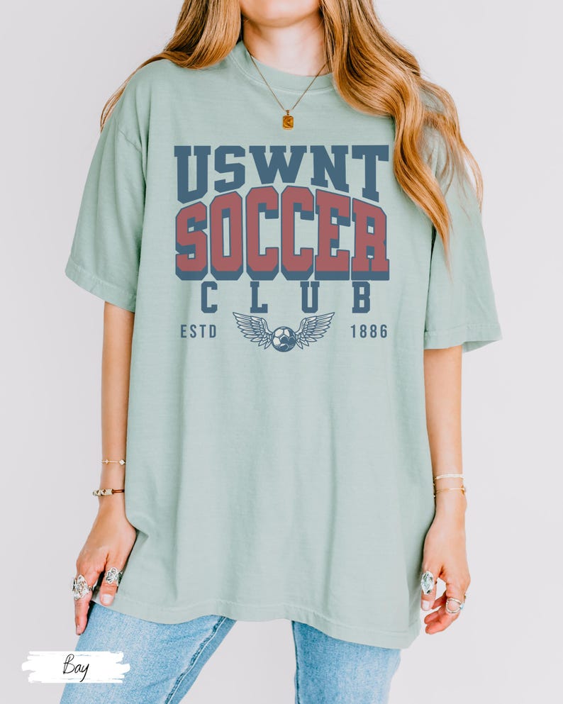 Puede incluir: Una camiseta de gran tama&ntilde;o de color verde claro con un dise&ntilde;o gr&aacute;fico de estilo vintage que dice "USWNT SOCCER CLUB ESTD 1886". El gr&aacute;fico presenta un bal&oacute;n de f&uacute;tbol con alas.