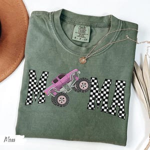 May include: Moss green t-shirt with the word "MAMA" in a black and white checkered pattern, featuring a pink monster truck graphic. The shirt has a crew neck and short sleeves. A gold necklace with a pendant is on the shirt.