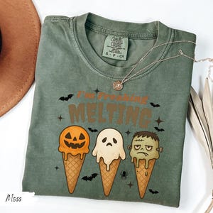May include: Moss green t-shirt with the text "I'm Freaking Melting" and illustrations of Halloween-themed ice cream cones. The cones feature a jack-o'-lantern, a ghost, and Frankenstein's monster. A silver necklace is draped across the shirt.