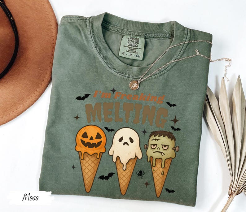 May include: Moss green t-shirt with the text "I'm Freaking Melting" and illustrations of Halloween-themed ice cream cones. The cones feature a jack-o'-lantern, a ghost, and Frankenstein's monster. A silver necklace is draped across the shirt.