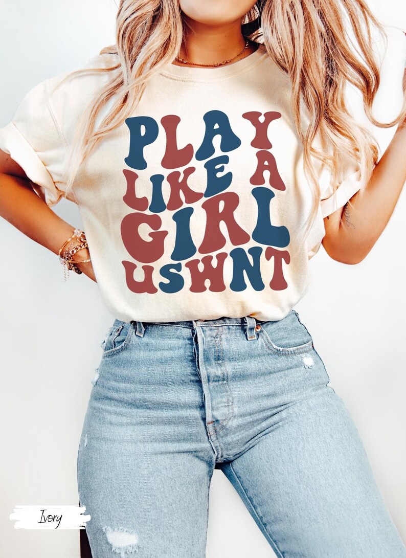 May include: A white t-shirt with the text "PLAY LIKE A GIRL USWNT" in blue and red retro font.