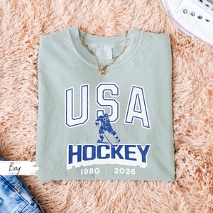 May include: A sage green crew neck sweatshirt with the text "USA HOCKEY" in white and blue. The design includes a hockey player graphic and the years 1980 and 2026. A gold necklace is draped on the shirt.