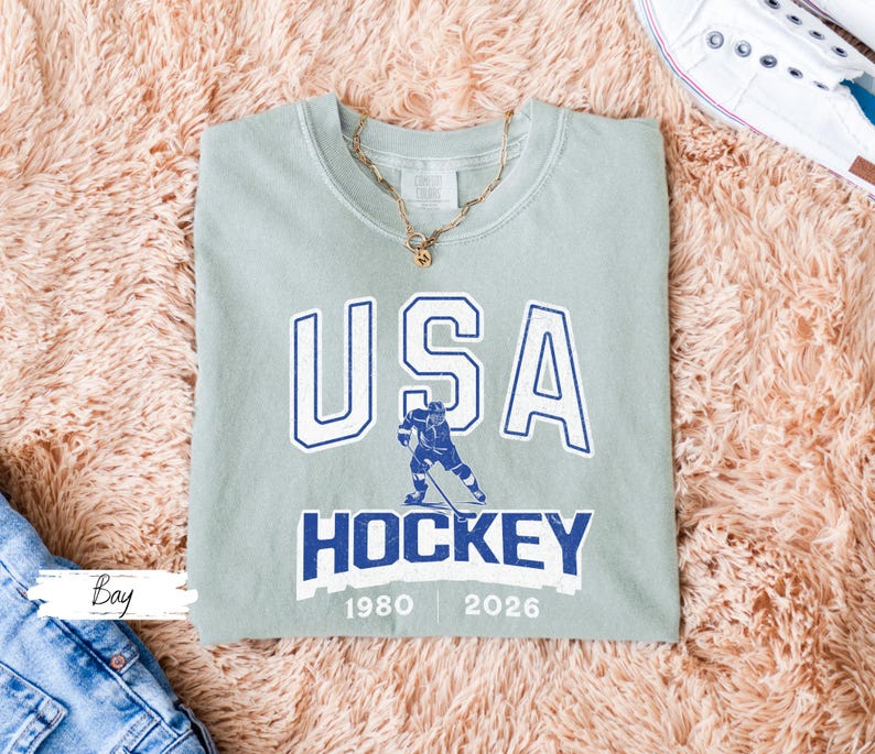 May include: A sage green crew neck sweatshirt with the text "USA HOCKEY" in white and blue. The design includes a hockey player graphic and the years 1980 and 2026. A gold necklace is draped on the shirt.