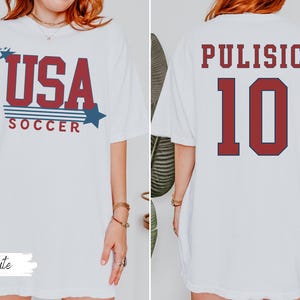 May include: White t-shirt with "USA SOCCER" in red and blue text on the front. The back of the shirt displays "PULISIC" and the number "10" in red with a navy blue outline. A casual, oversized fit.