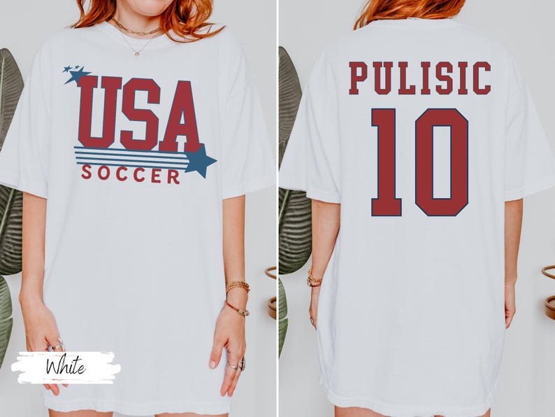 May include: White t-shirt with "USA SOCCER" in red and blue text on the front. The back of the shirt displays "PULISIC" and the number "10" in red with a navy blue outline. A casual, oversized fit.