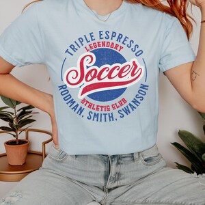 Puede incluir: Una camiseta azul claro con un dise&ntilde;o gr&aacute;fico de estilo vintage. El gr&aacute;fico presenta un c&iacute;rculo azul y rojo con la palabra "Soccer" en una fuente de letra roja y blanca. El c&iacute;rculo tambi&eacute;n incluye el texto "Triple Espresso Legendary Athletic Club Rodman, Smith, Swanson".