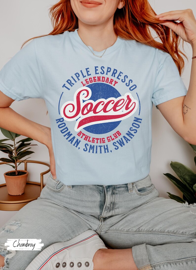 Puede incluir: Una camiseta azul claro con un dise&ntilde;o gr&aacute;fico de estilo vintage. El gr&aacute;fico presenta un c&iacute;rculo azul y rojo con la palabra "Soccer" en una fuente de letra roja y blanca. El c&iacute;rculo tambi&eacute;n incluye el texto "Triple Espresso Legendary Athletic Club Rodman, Smith, Swanson".