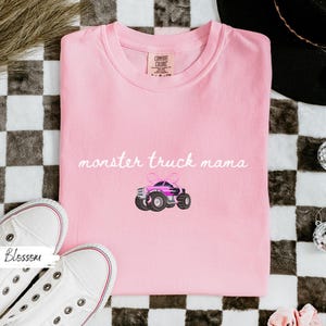 Puede incluir: Una camiseta Comfort Colors rosa claro con la frase "monster truck mama" en cursiva. Debajo, un dibujo de un monster truck rosa y negro con un lazo. Zapatillas blancas en la esquina inferior izquierda.