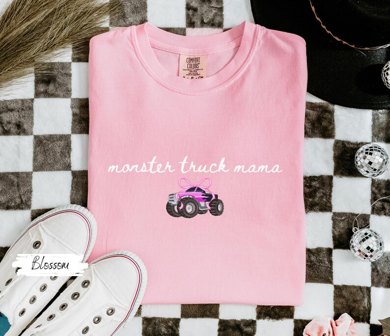 Puede incluir: Una camiseta Comfort Colors rosa claro con la frase "monster truck mama" en cursiva. Debajo, un dibujo de un monster truck rosa y negro con un lazo. Zapatillas blancas en la esquina inferior izquierda.