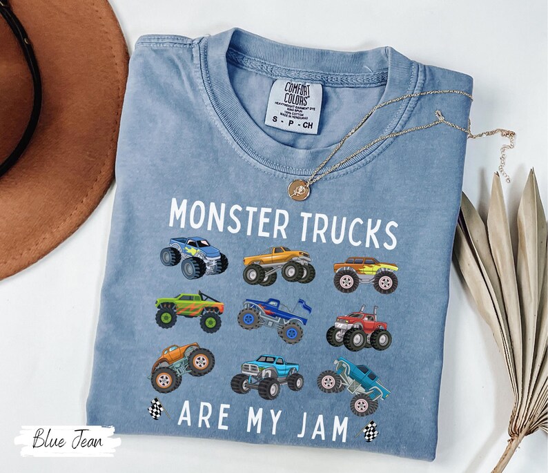 May include: A light blue t-shirt featuring the text "MONSTER TRUCKS ARE MY JAM" and illustrations of monster trucks. The shirt has a crew neck and is made of a soft material.