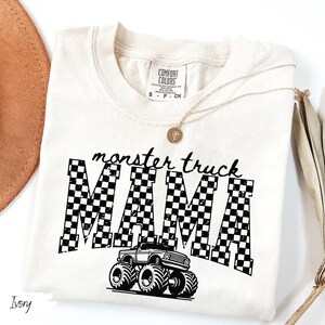 Op de afbeelding: Ivoor t-shirt met de zwarte tekst "monster truck MAMA". Het woord "MAMA" heeft een zwart-wit geblokt patroon, met een afbeelding van een monstertruck eronder. Een gouden ketting en een bruine hoed zijn ook zichtbaar.