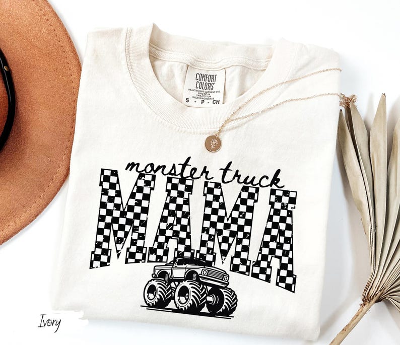 Op de afbeelding: Ivoor t-shirt met de zwarte tekst "monster truck MAMA". Het woord "MAMA" heeft een zwart-wit geblokt patroon, met een afbeelding van een monstertruck eronder. Een gouden ketting en een bruine hoed zijn ook zichtbaar.