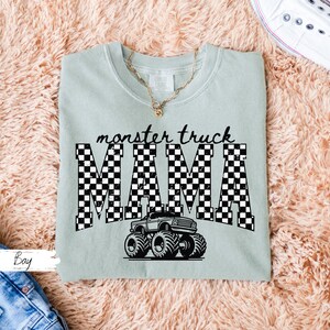 Op de afbeelding: Lichtgroen T-shirt met de tekst "monster truck MAMA" in zwart-witte geblokte letters. Een zwart-witte monster truck afbeelding staat eronder. Het shirt ligt op een roze, pluizig oppervlak.