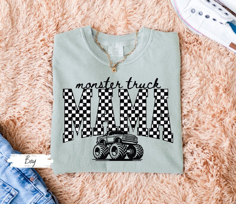 Op de afbeelding: Lichtgroen T-shirt met de tekst "monster truck MAMA" in zwart-witte geblokte letters. Een zwart-witte monster truck afbeelding staat eronder. Het shirt ligt op een roze, pluizig oppervlak.