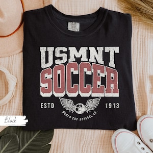 Może przedstawiać: Czarna koszulka z wytartą biało-czerwoną grafiką z napisem "USMNT SOCCER ESTD 1913" z piłką nożną i wzorem skrzydeł. Koszulka wykonana jest z miękkiego materiału.