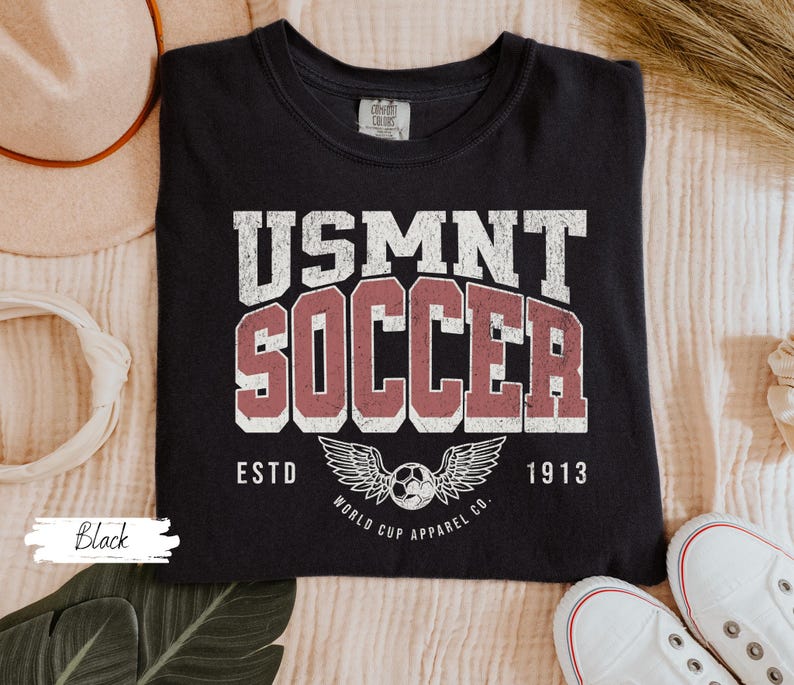 Może przedstawiać: Czarna koszulka z wytartą biało-czerwoną grafiką z napisem "USMNT SOCCER ESTD 1913" z piłką nożną i wzorem skrzydeł. Koszulka wykonana jest z miękkiego materiału.