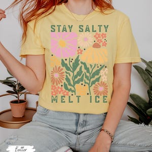 Op de afbeelding: Een geel t-shirt met de tekst "STAY SALTY MELT ICE" in het groen. Het shirt heeft een bloemenontwerp met roze, oranje en gele bloemen en groene bladeren. Het shirt is gemaakt van een zacht materiaal.