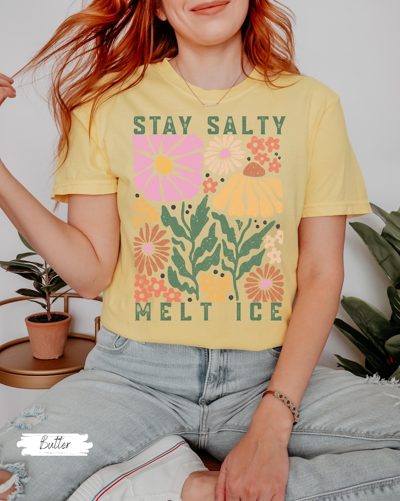 Op de afbeelding: Een geel t-shirt met de tekst "STAY SALTY MELT ICE" in het groen. Het shirt heeft een bloemenontwerp met roze, oranje en gele bloemen en groene bladeren. Het shirt is gemaakt van een zacht materiaal.